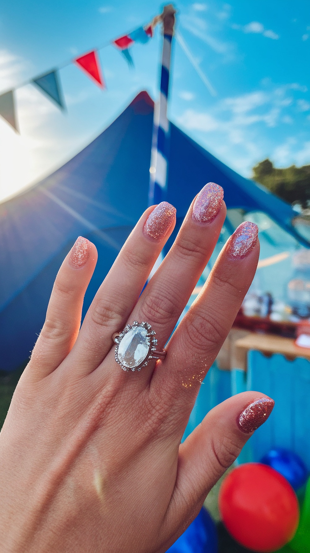 A hand with glittery pink and gold nails, showcasing a ring, against a colorful summer background.