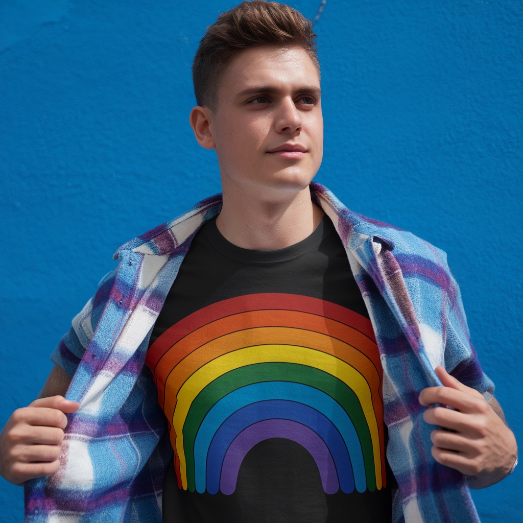 A young man wearing a rainbow pride t-shirt with a plaid shirt over it, standing against a blue wall.
