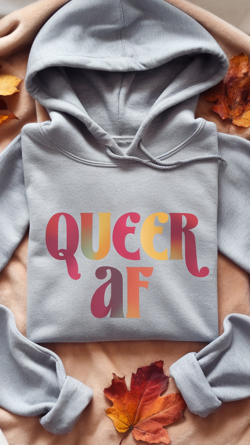 A close-up of a grey hoodie featuring the text 'Queer, af' in colorful letters, surrounded by autumn leaves.