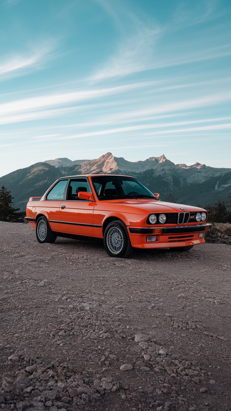 A vibrant orange BMW E30 318Is parked on a gravel road with mountains in the background.