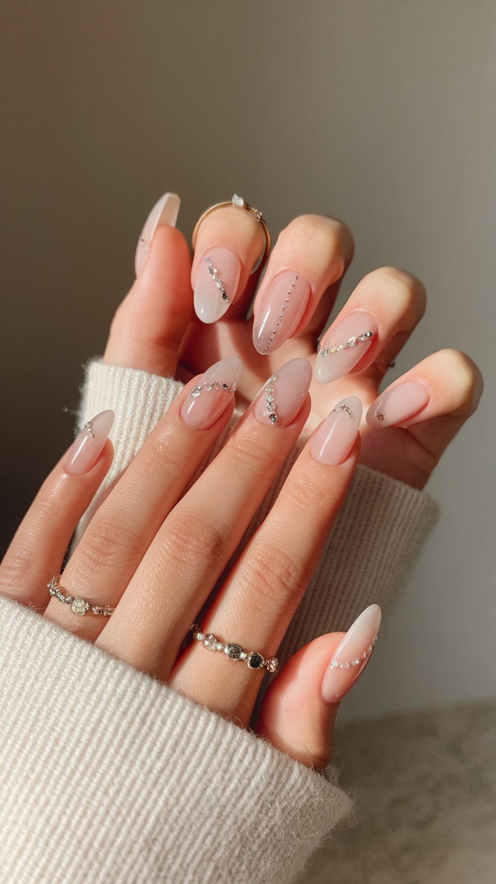 A close-up of hands with slim, almond-shaped nails adorned with rhinestones, wearing delicate rings.