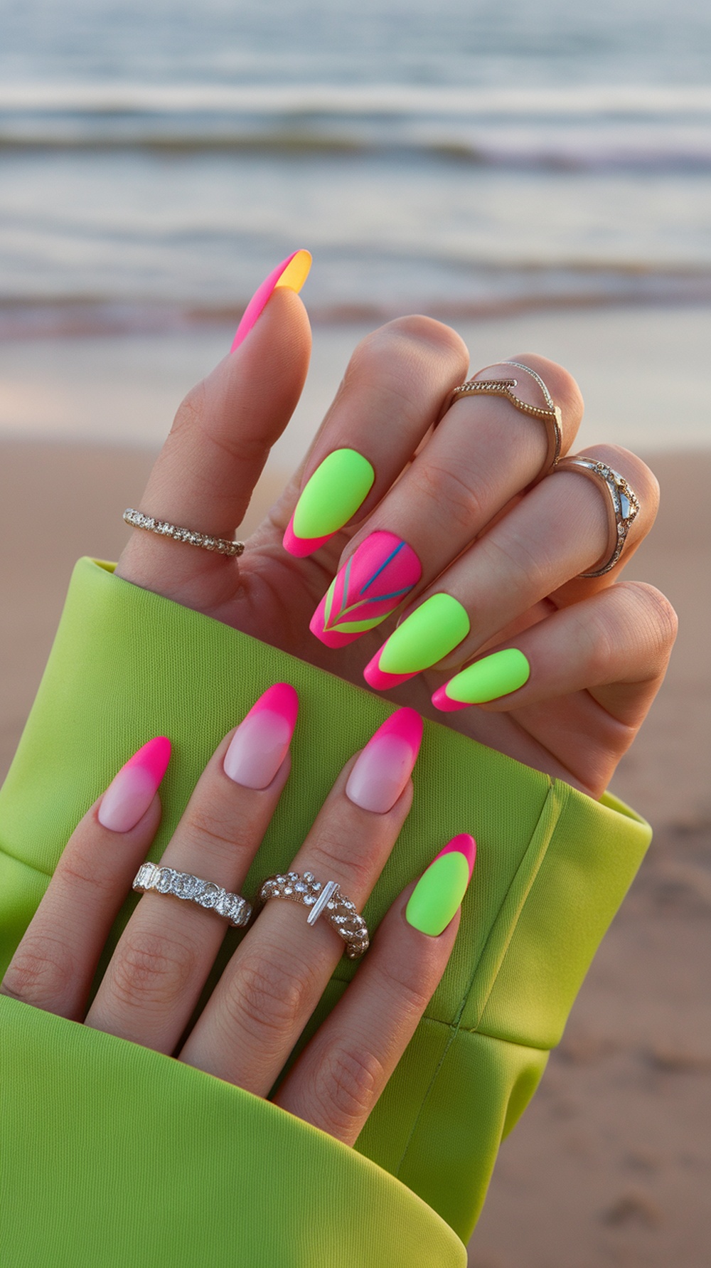 Close-up of hands with bright neon nails featuring pink and green colors, accessorized with rings, against a beach background.