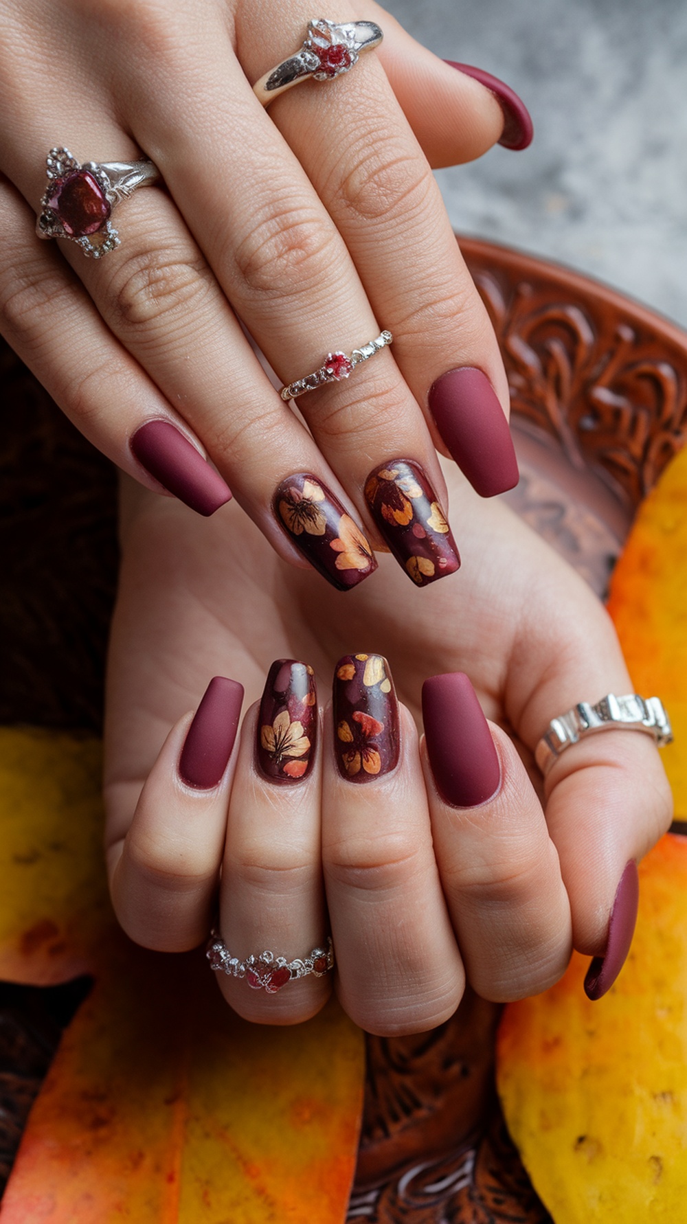 Nails painted in burgundy and brown with floral designs, complemented by silver rings.