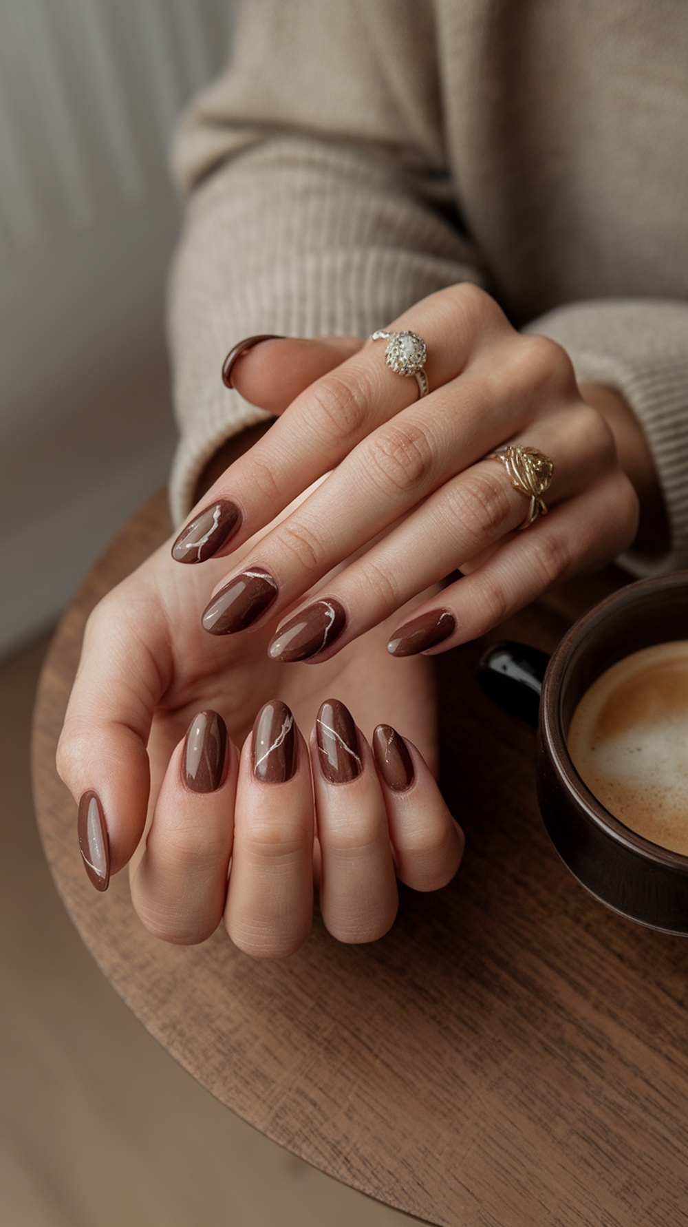A close-up of hands with chestnut brown nails featuring a marble effect, resting on a wooden table next to a cup of coffee.