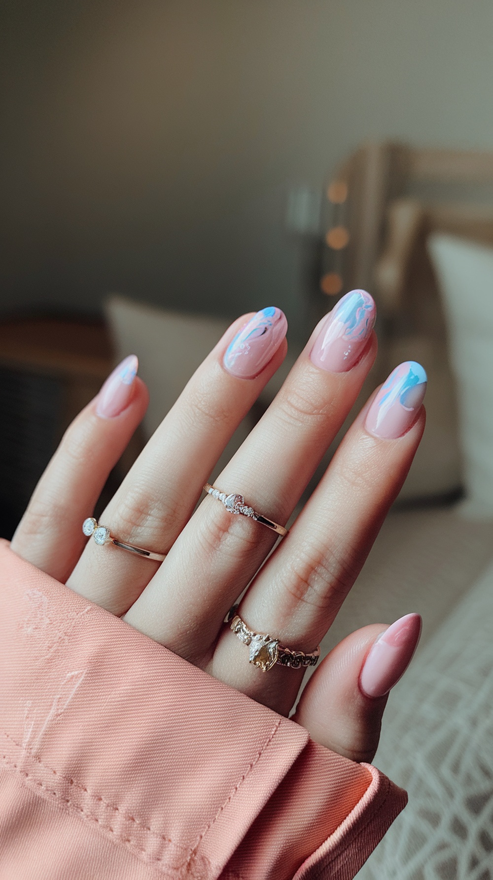 Close-up of hands with cotton candy chrome nails, featuring a soft pink base with blue accents, adorned with delicate rings.