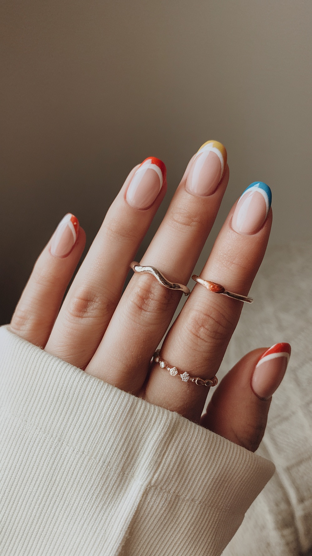 Nails with colorful French tips in red, yellow, and blue, featuring a nude base and delicate rings.