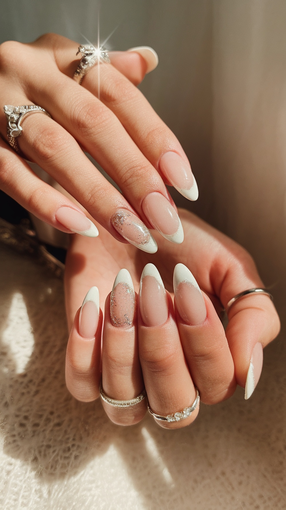 A hand with creamy white almond nails featuring subtle glitter, adorned with rings