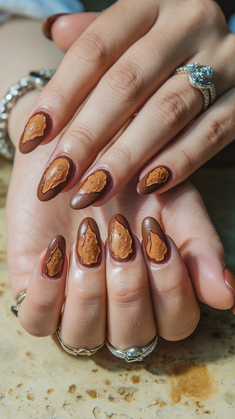 Close-up of hands with earthy brown nails featuring textured accents, adorned with rings.