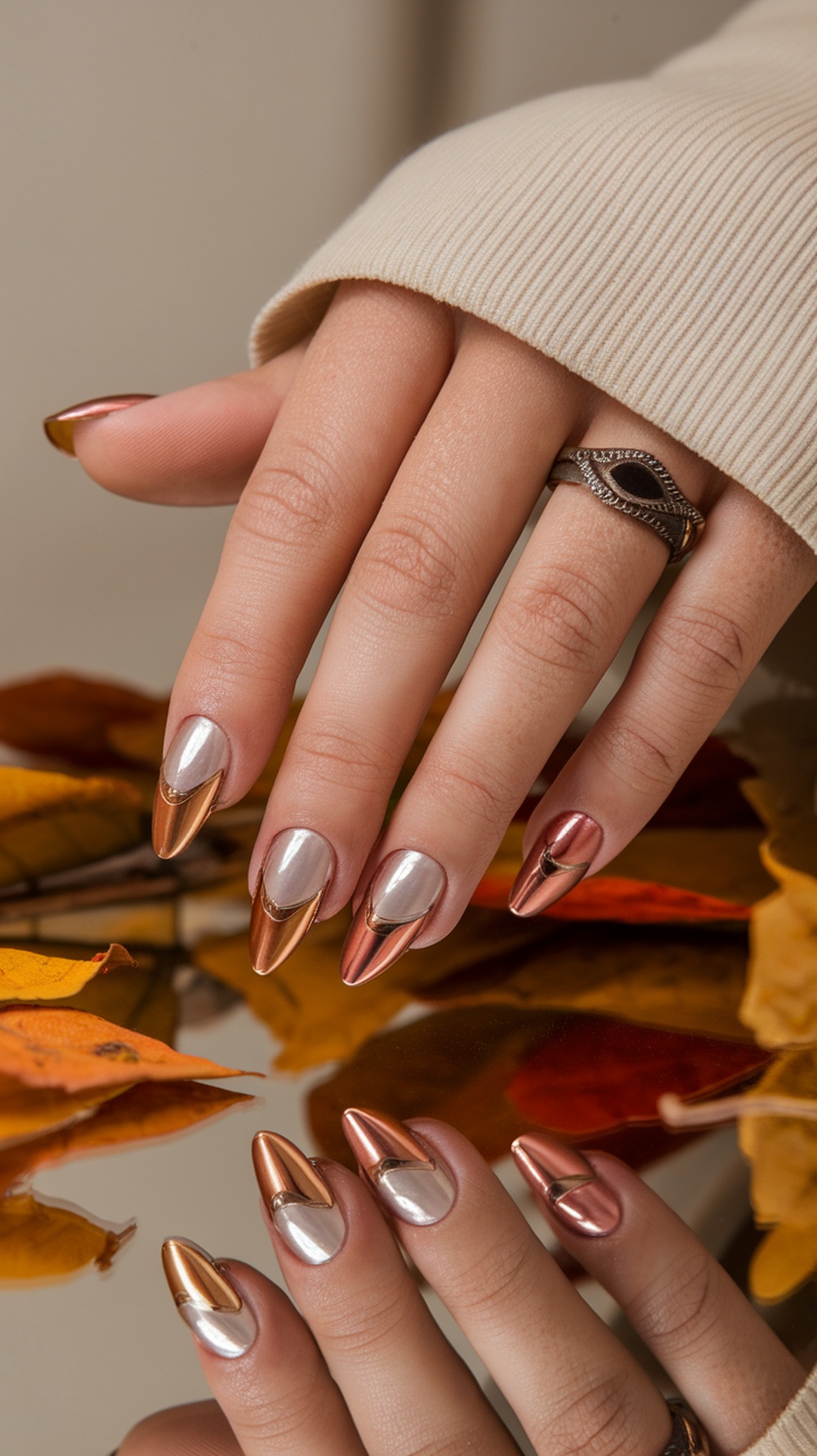 Close-up of elegantly designed nails with metallic copper and silver accents, surrounded by autumn leaves.