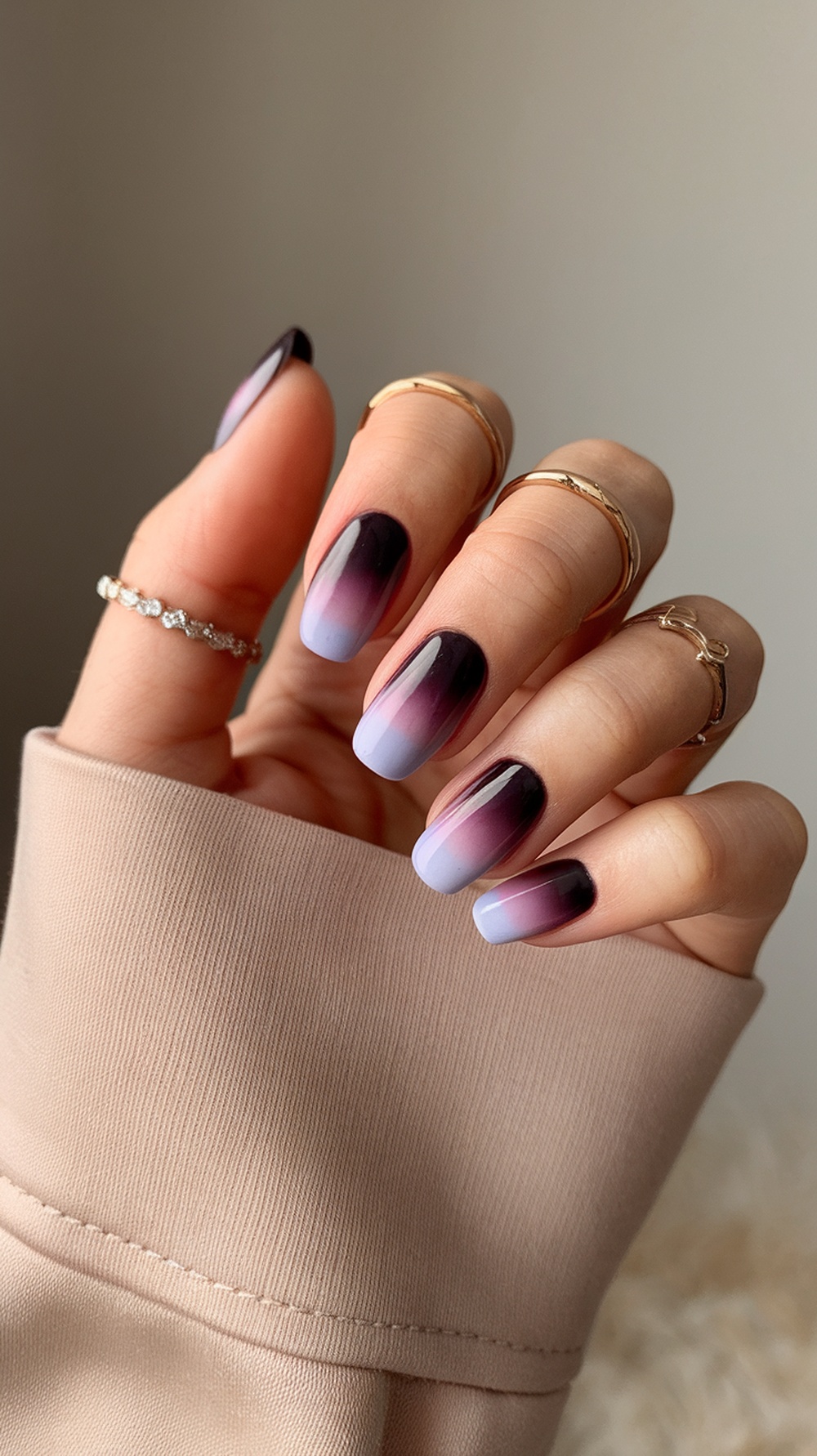 A close-up of elegant plum ombre nails transitioning from dark plum to light lavender, adorned with gold rings.