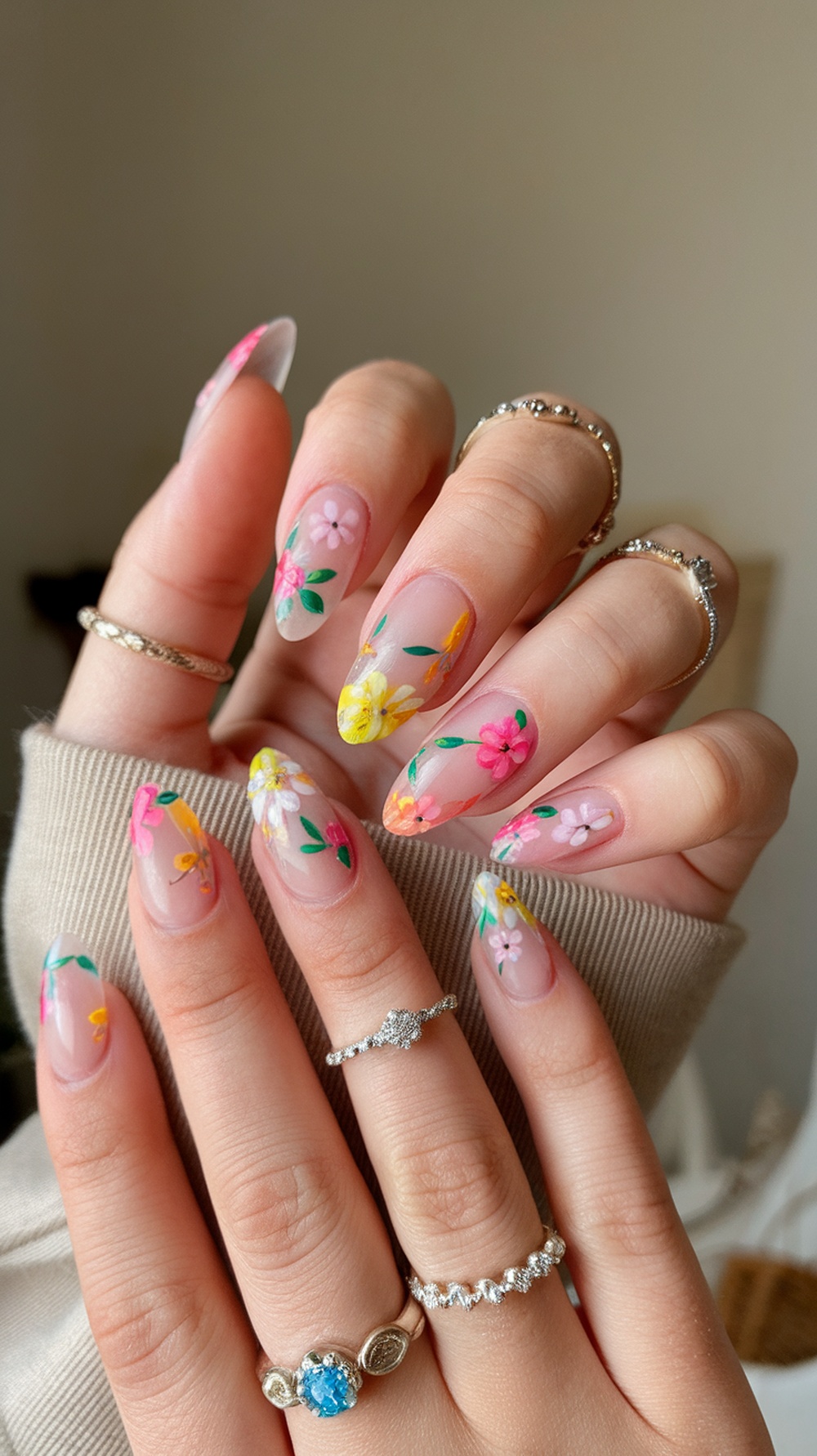 A close-up of hands with floral accent almond nails, featuring colorful flowers on a clear base, adorned with rings.