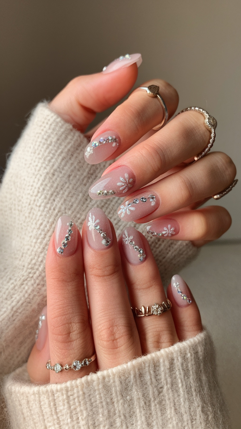 A close-up of hands with floral nail art featuring white flowers and rhinestones on a nude background.