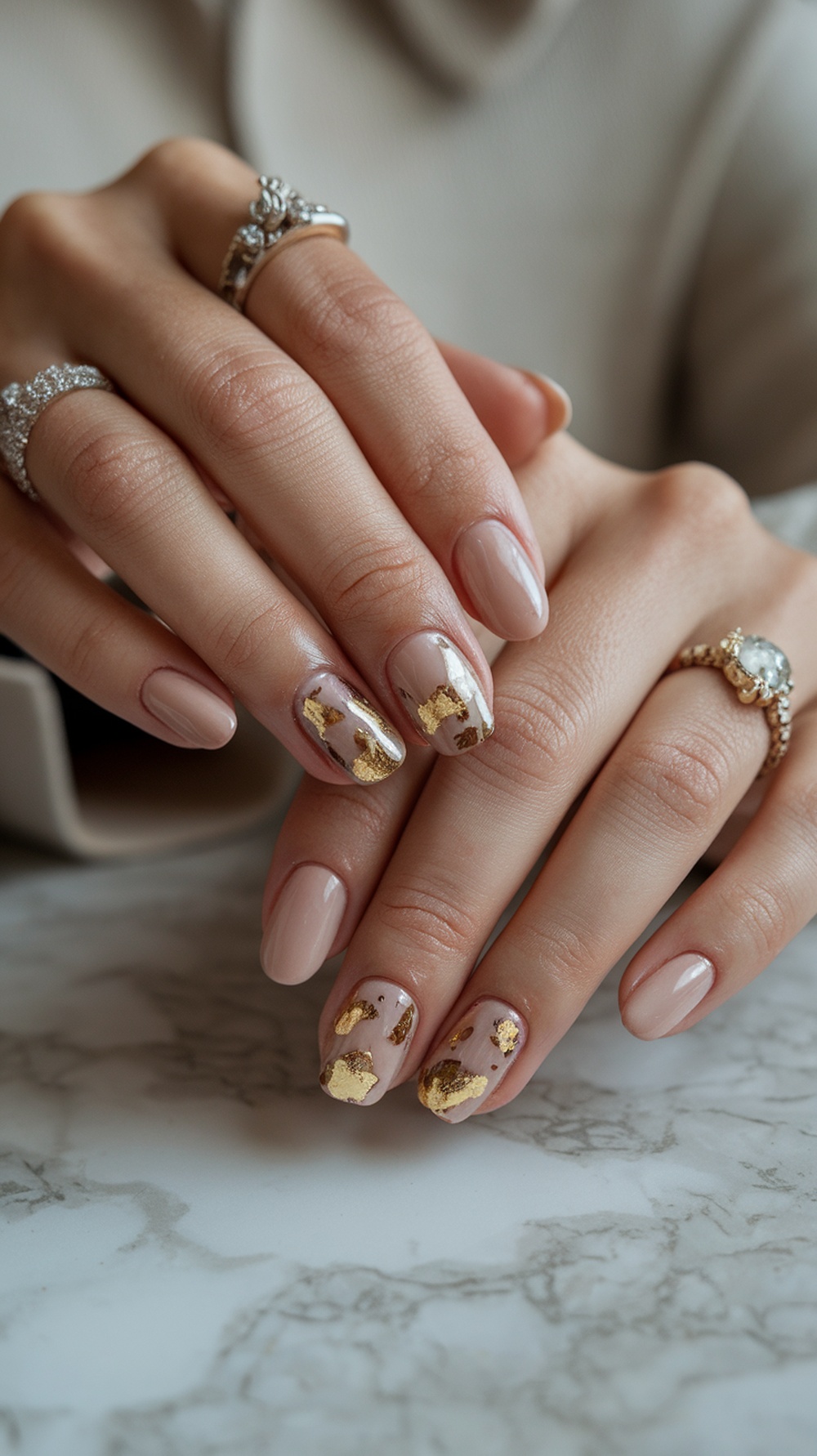 Close-up of hands with glossy nude nails featuring gold foil accents, adorned with rings.