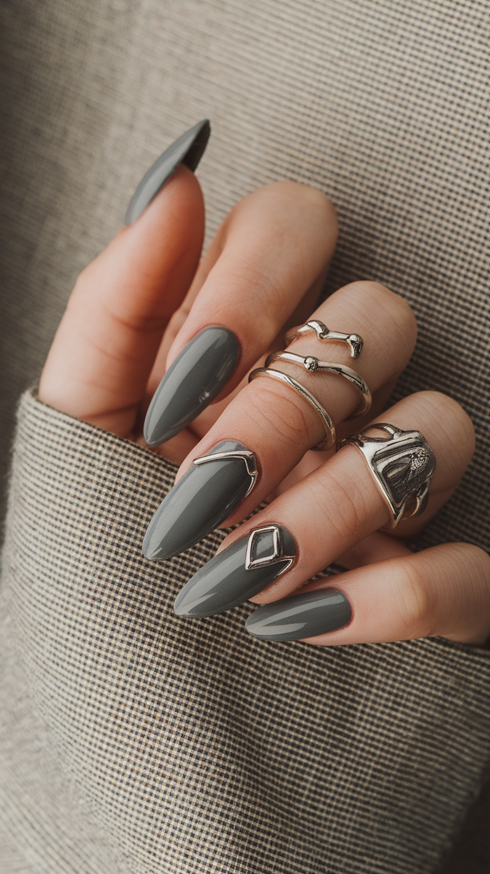 A close-up of hands with light charcoal almond nails and silver rings against a textured background.