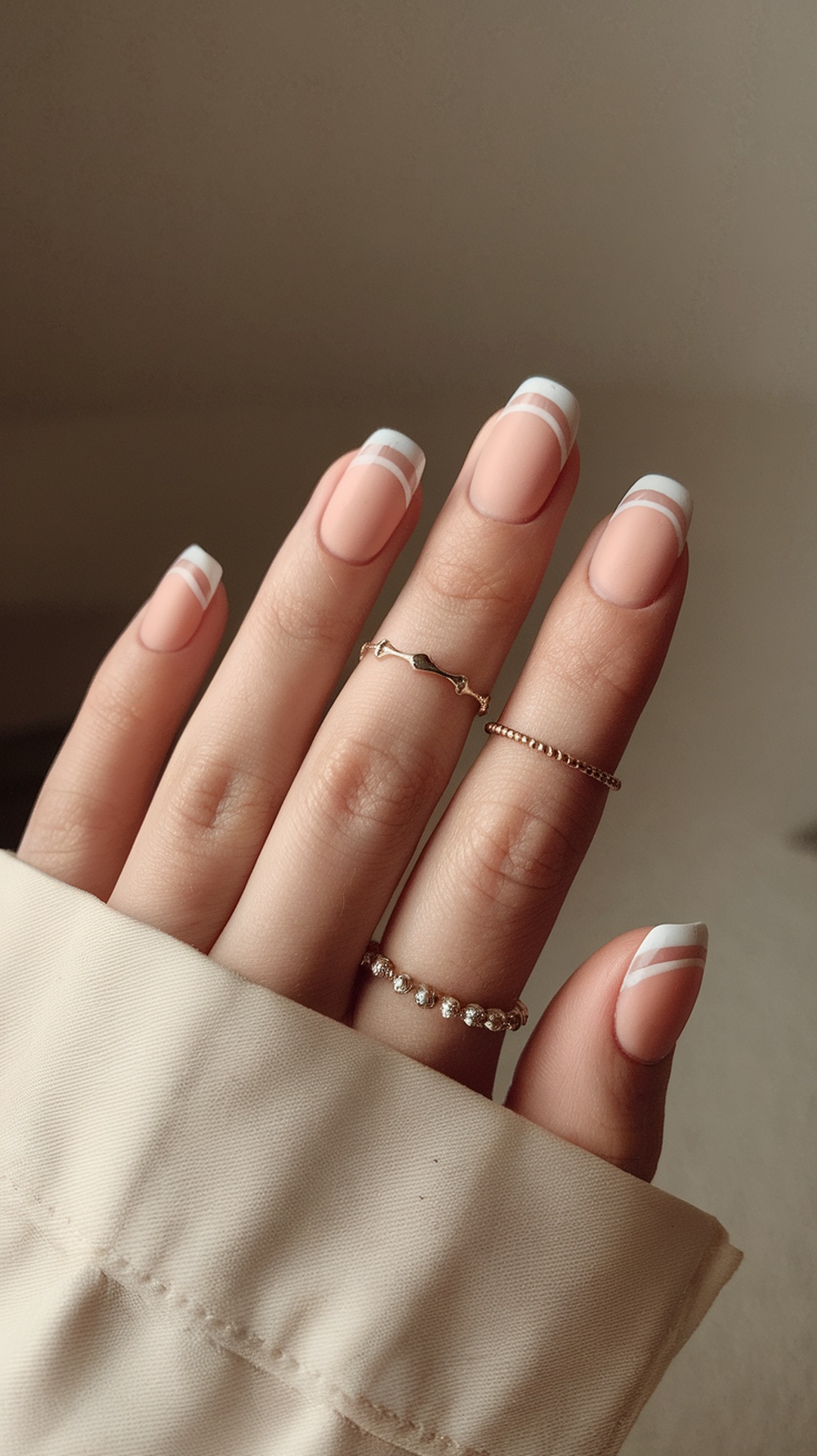 A close-up of hands with matte finish French nails, featuring a nude base and white tips, adorned with gold rings.