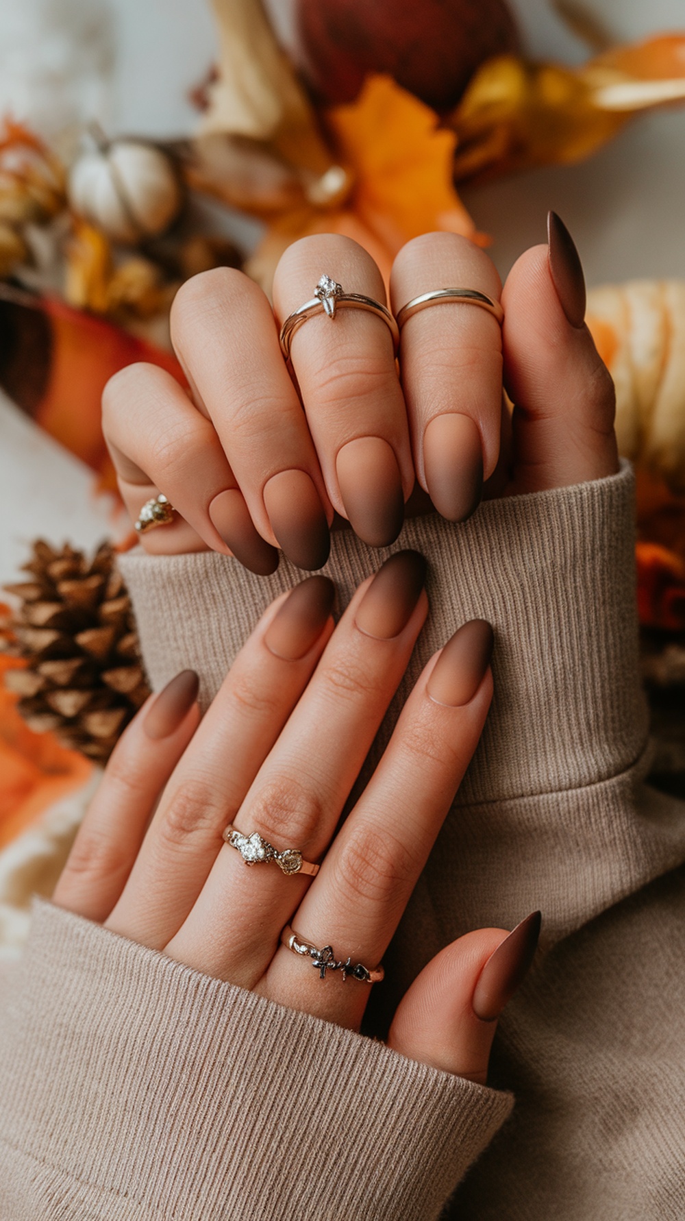 Close-up of hands with matte mocha ombre nails and gold rings, surrounded by fall decorations.