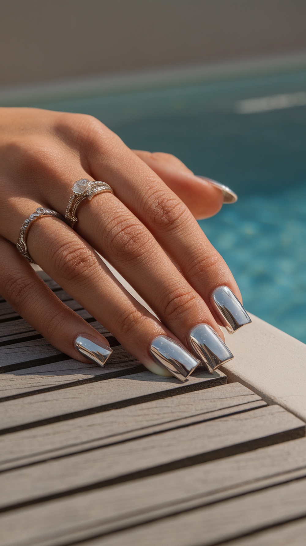 Close-up of a hand with metallic square nails resting on a wooden surface, with a pool in the background.