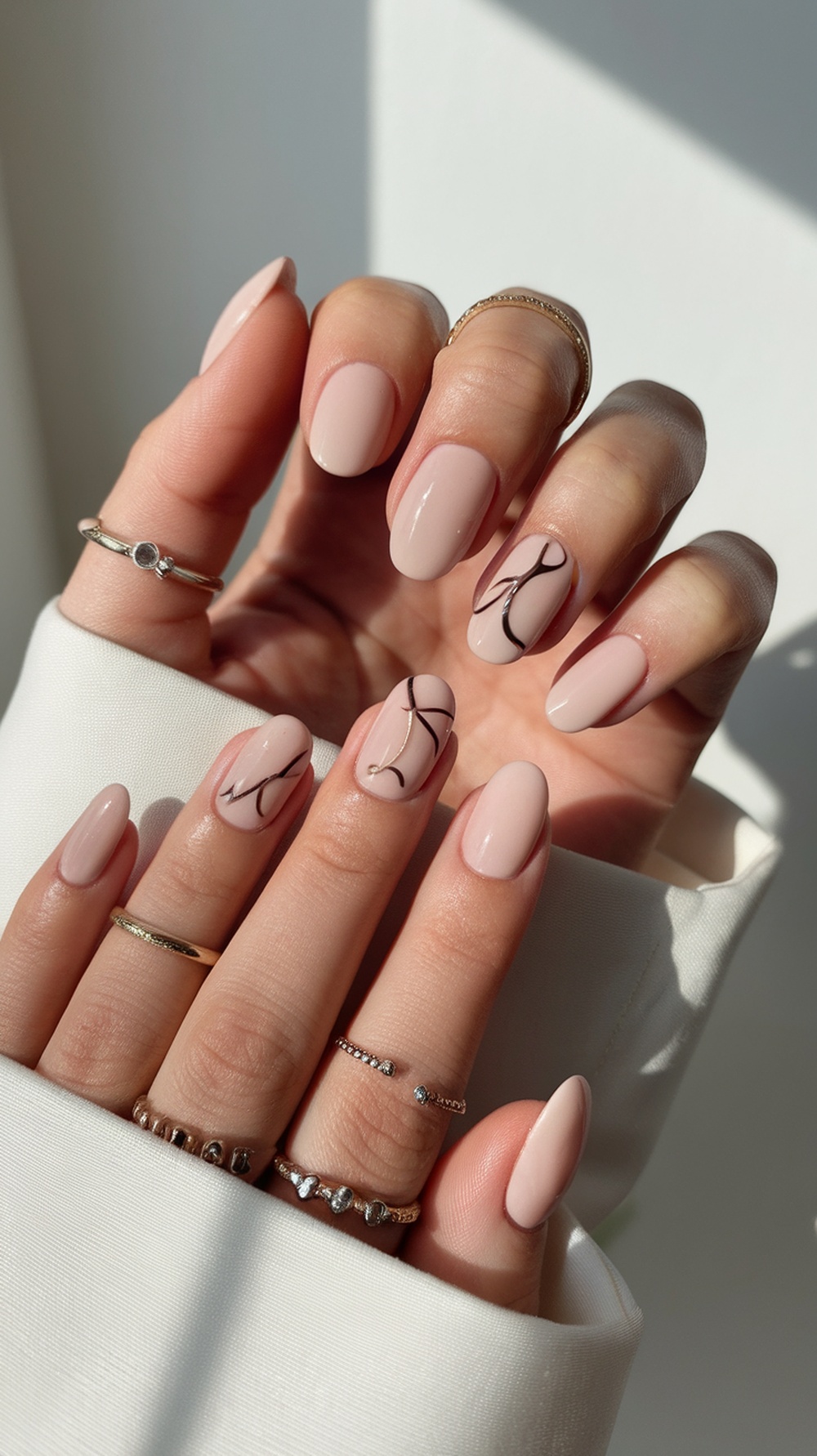 Close-up of hands with nude fall nails featuring minimalist black line art and multiple rings.