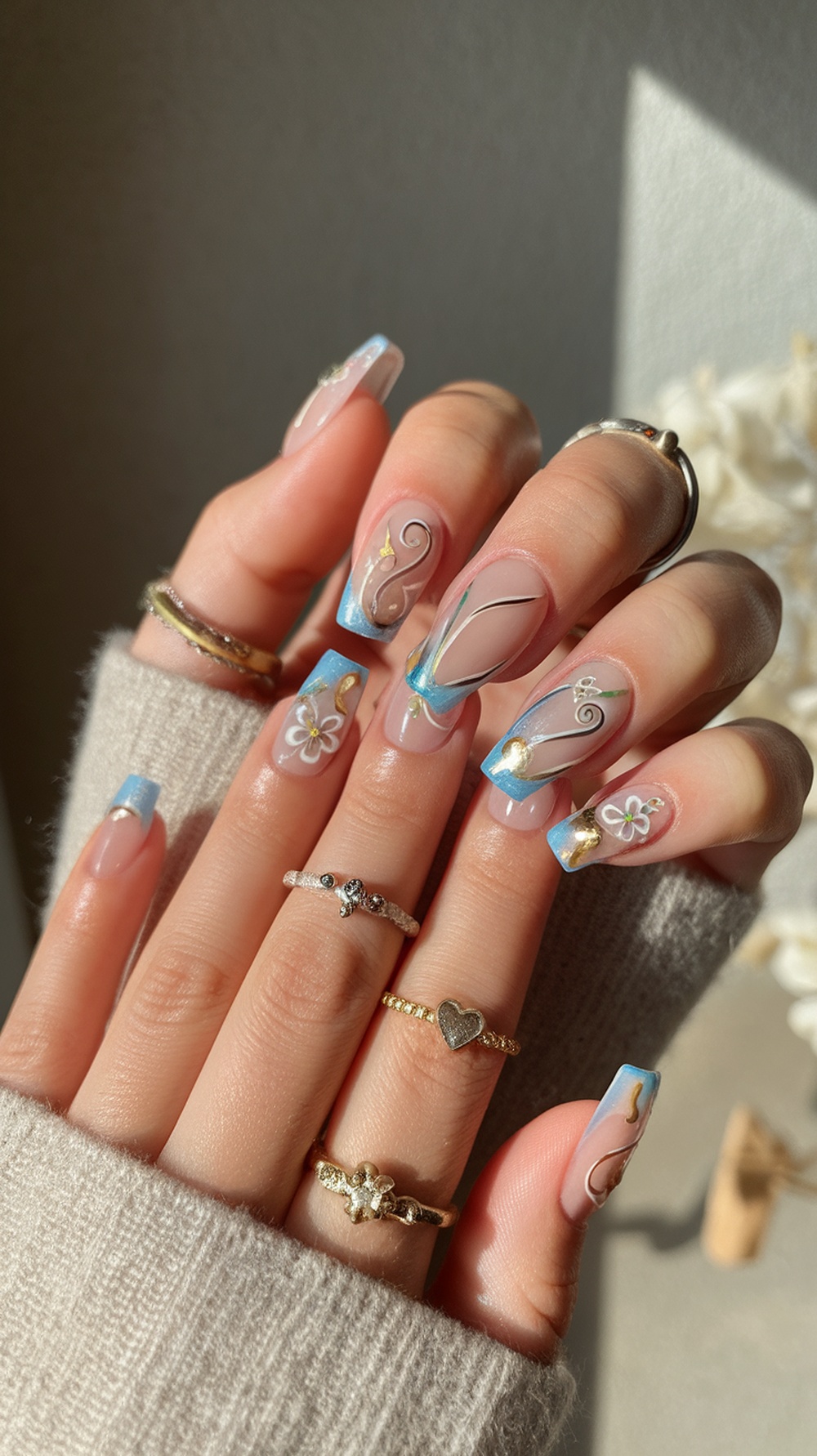 A close-up of tapered square nails with blue tips and intricate gold and floral designs, accessorized with rings.