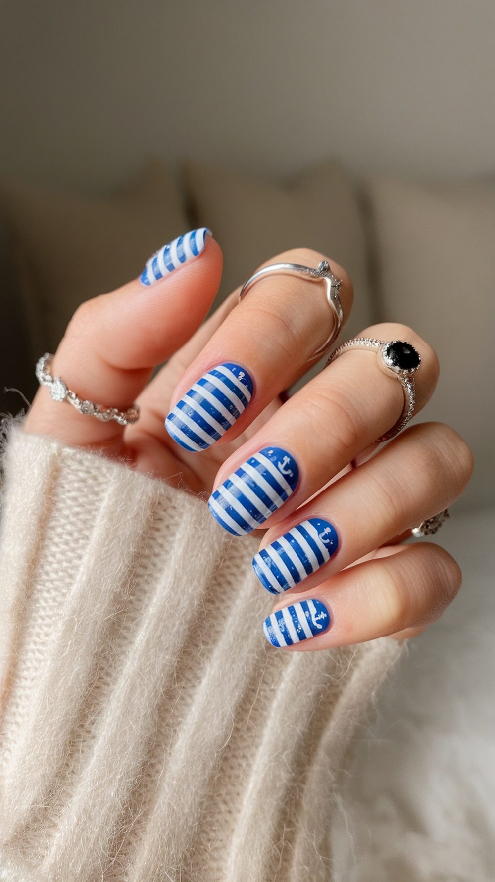 Nail design featuring blue and white stripes with small anchors, complemented by silver rings and a cozy sweater.