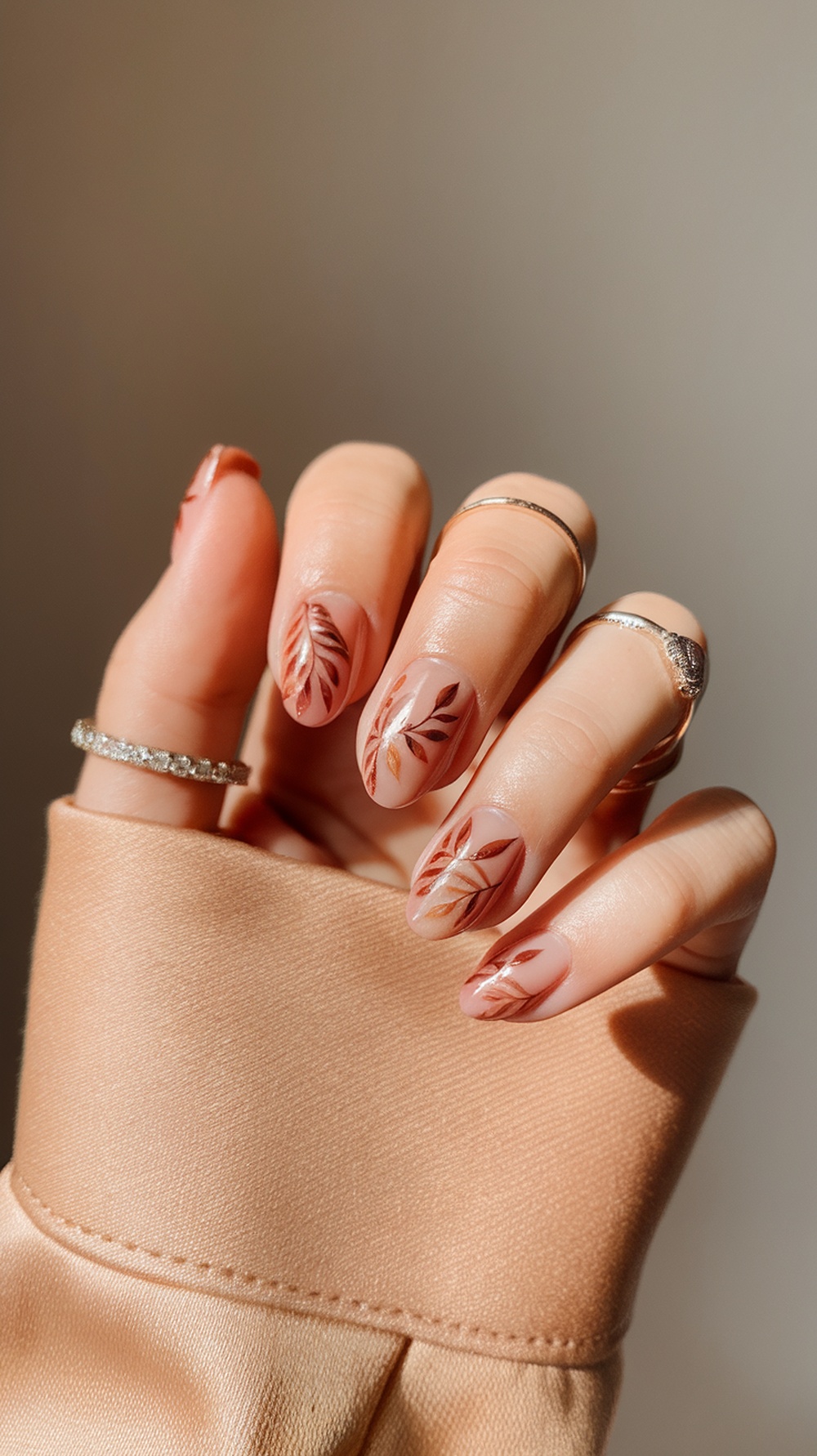 Nude nails with leaf patterns in earthy tones, accessorized with rings, on a soft fabric background.