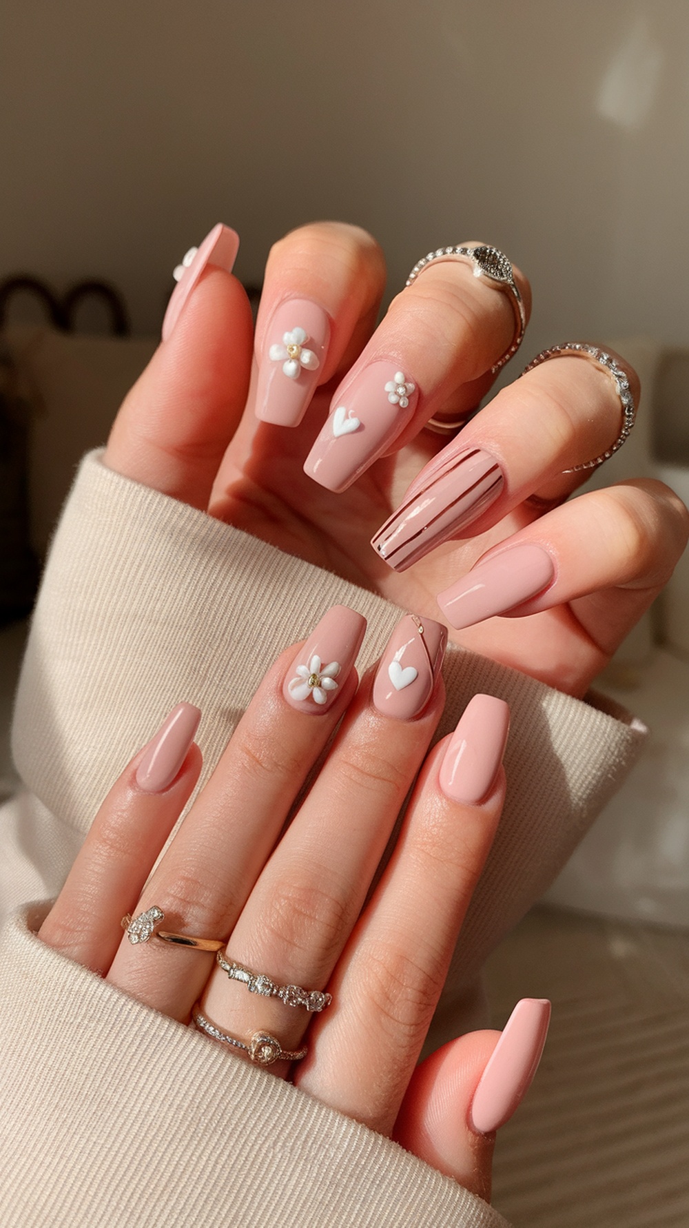 Nude pink nails with floral and heart designs, accessorized with rings