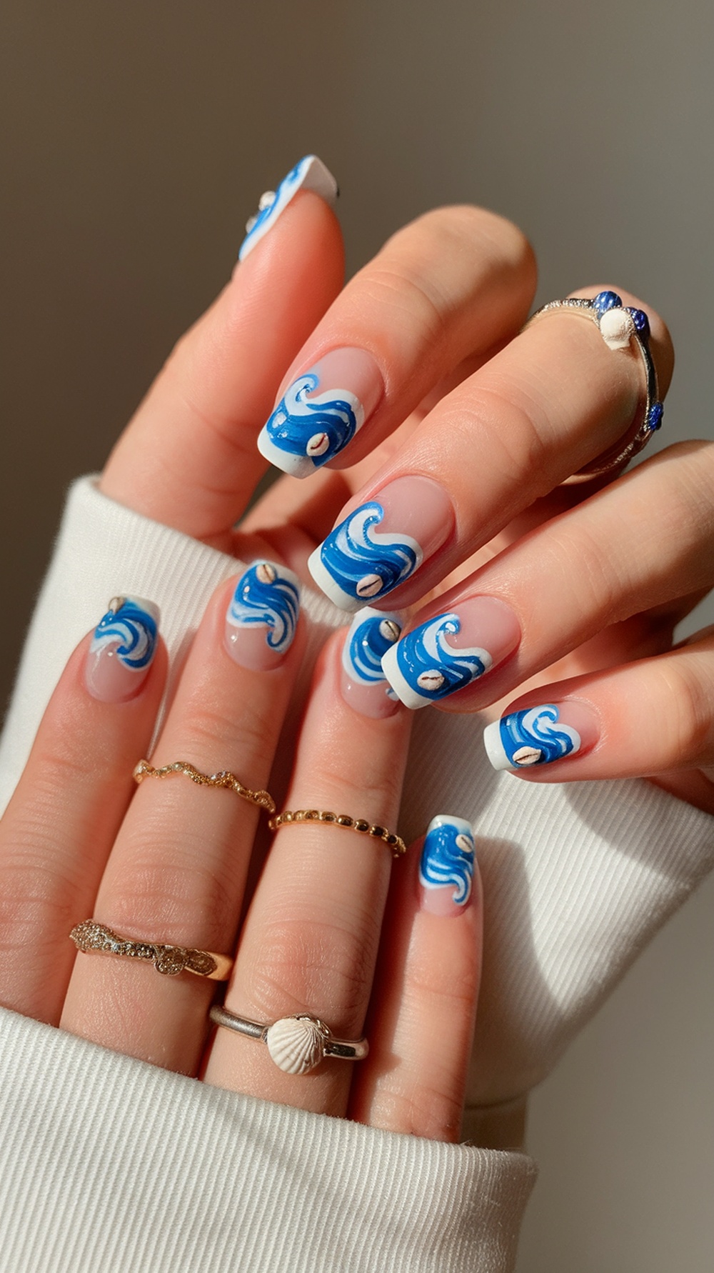 Close-up of hands with ocean wave nail art featuring blue and white designs, adorned with rings.