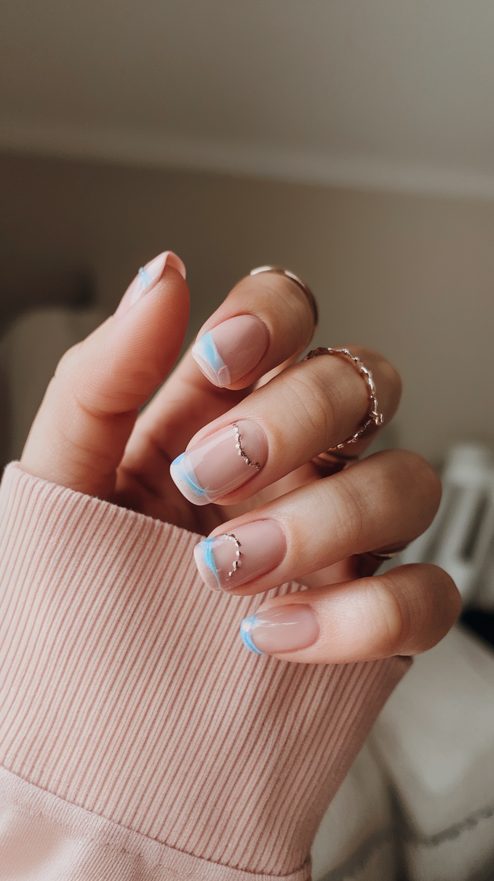 A close-up of a hand with pastel French tip nails featuring light blue tips and delicate chain designs, resting on a soft pink sweater.