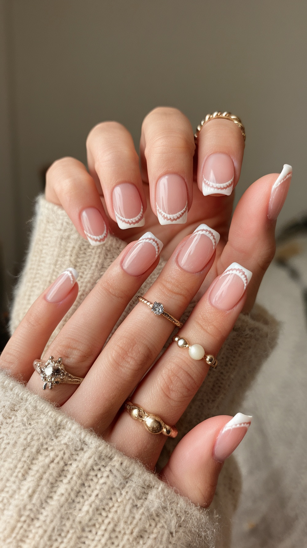 Close-up of hands with seashell French tip nails, featuring a soft pink base and white scalloped tips, adorned with rings.