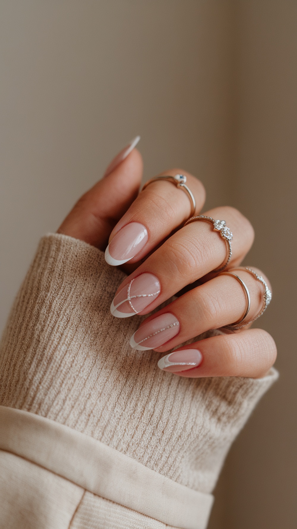 A hand with nude pink nails featuring a French tip and delicate designs, adorned with rings, resting on a beige sweater.