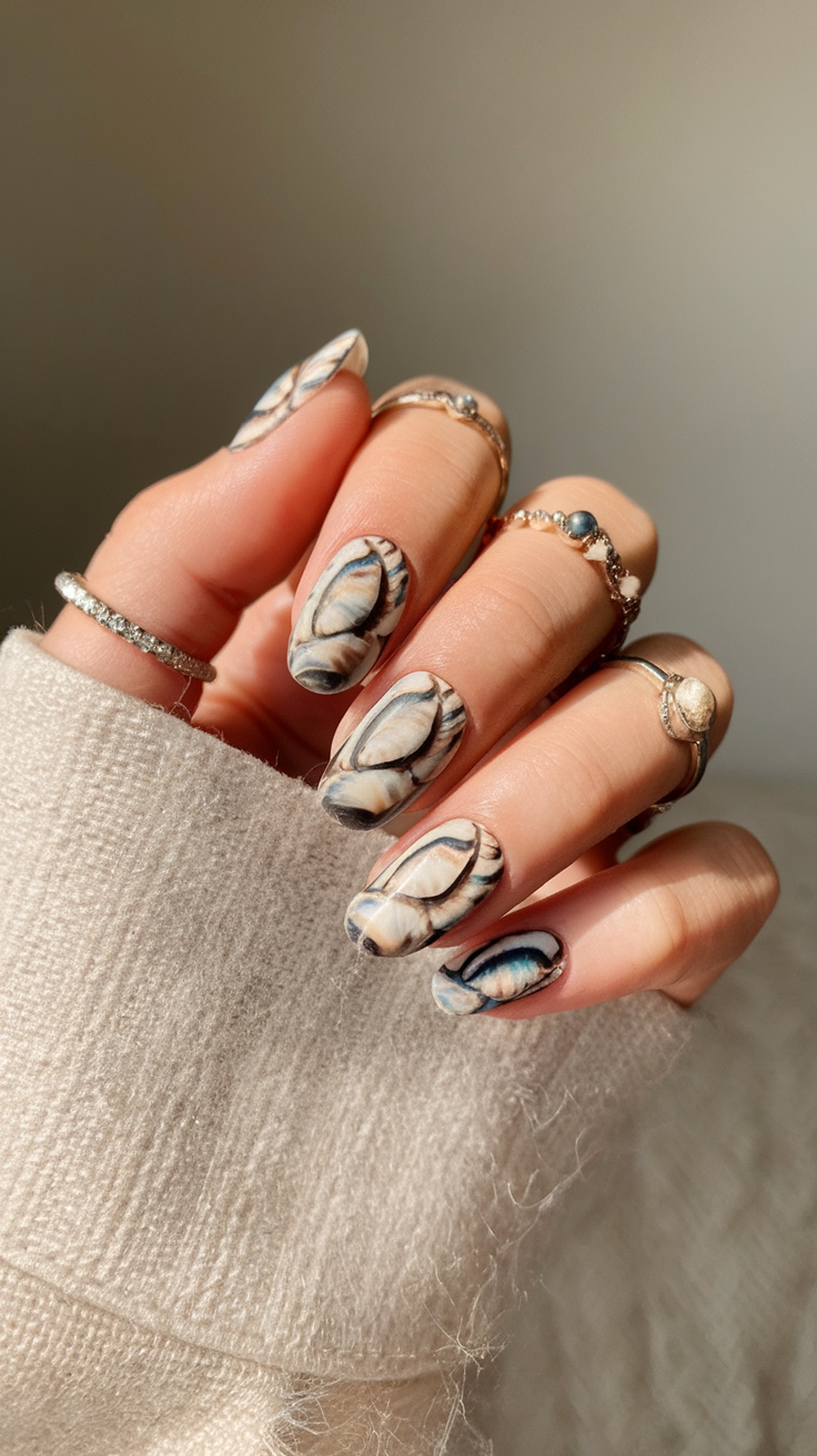 Close-up of shell-inspired nail art with intricate designs in beige, cream, and blue, paired with delicate rings.