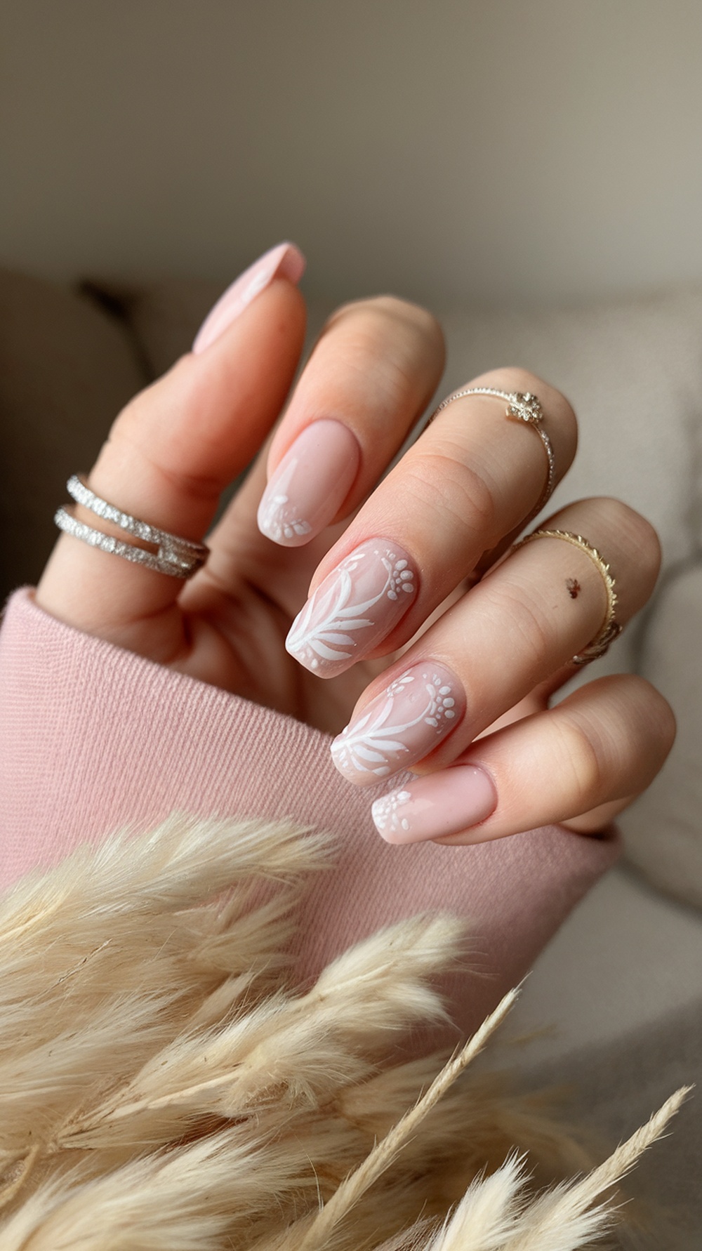 A hand showing soft pink nails with floral designs, adorned with rings, resting on a soft background.