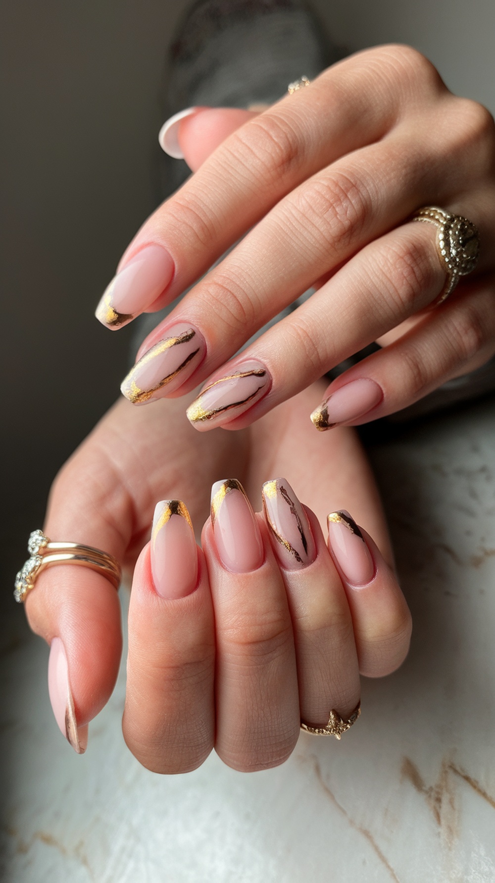 A close-up of hands with soft pink nails featuring gold accents and rings.