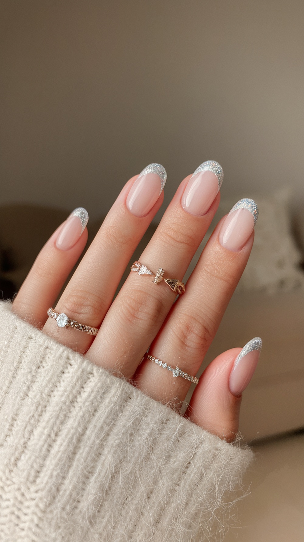 A close-up of hands with sparkling French tip nails and delicate rings, showcasing a nude base with silver glitter tips.