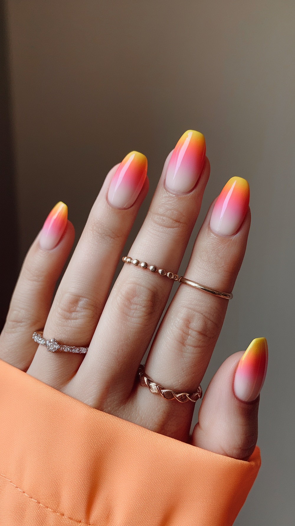 Close-up of hands with sunset ombre nails featuring pink and yellow gradient, adorned with rings and wearing an orange top.
