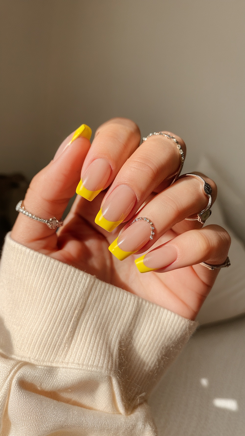 A hand with nails featuring sunshine yellow tips, adorned with rings, against a neutral background.