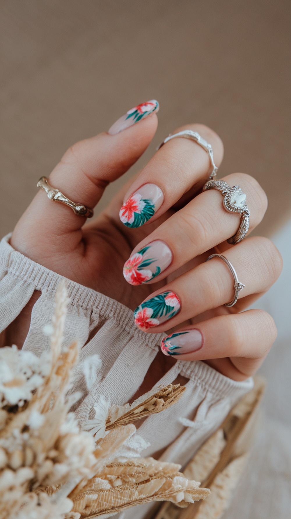 Close-up of short nails with tropical floral designs in red and green, complemented by delicate rings.