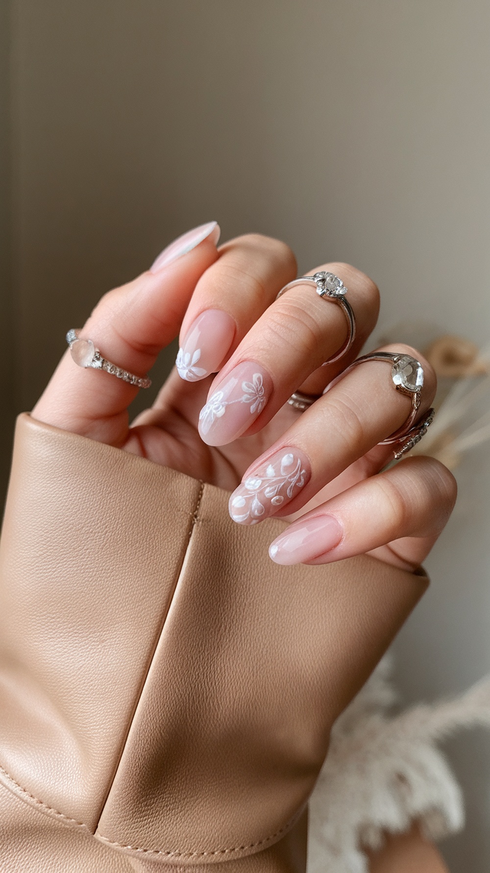 Close-up of a hand with tapered square nails featuring floral designs, wearing multiple rings, and resting on a beige leather surface.