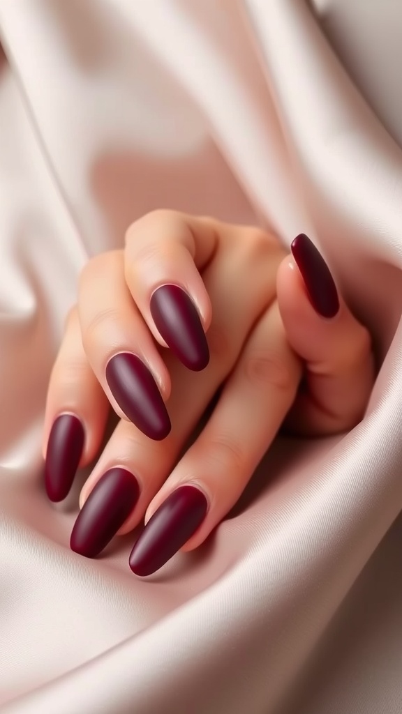 Close-up of hands with matte finish almond nails in deep burgundy color on a soft satin background.