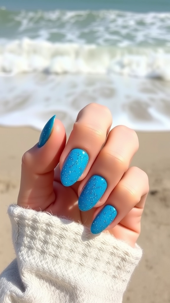 Close-up of ocean blue almond nails with glitter on a beach