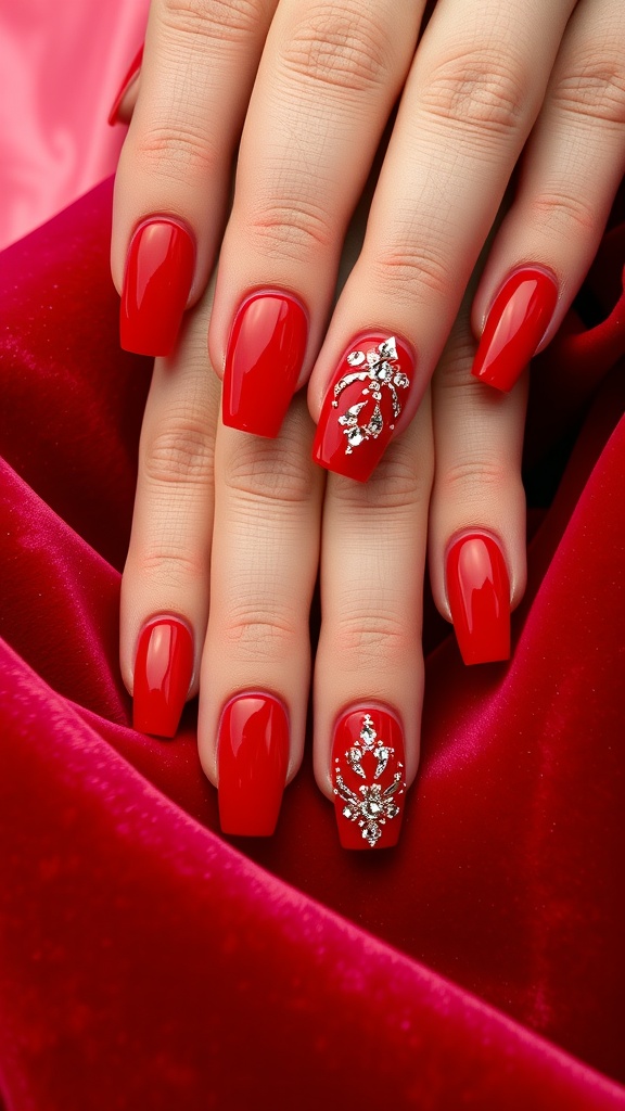 Close-up of hands with vibrant red nail polish and rhinestones on one nail, displayed on a soft red fabric background.
