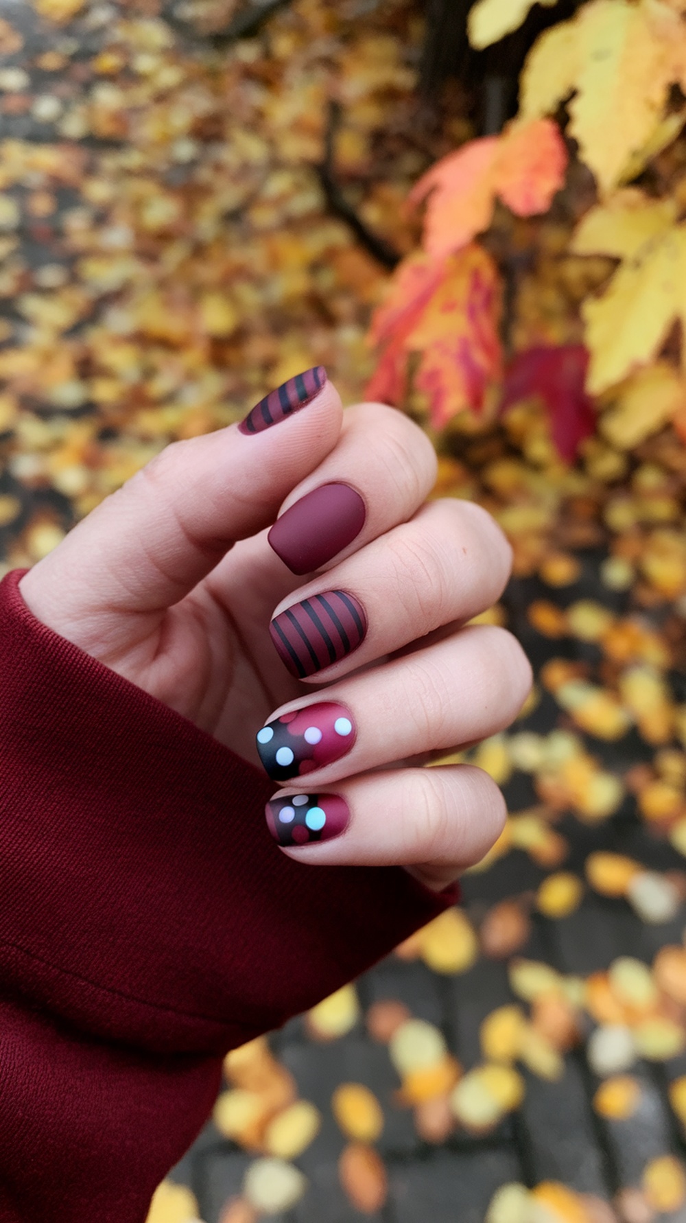 A hand showing burgundy nails with matte black stripes and colorful dots against a backdrop of autumn leaves.