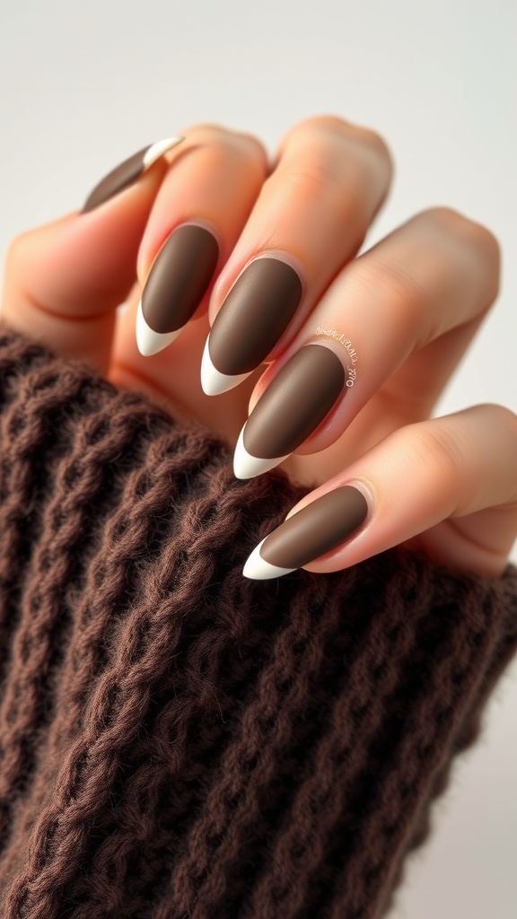 Almond-shaped nails with a matte cocoa finish and thin white tips, resting on a knitted brown sweater.