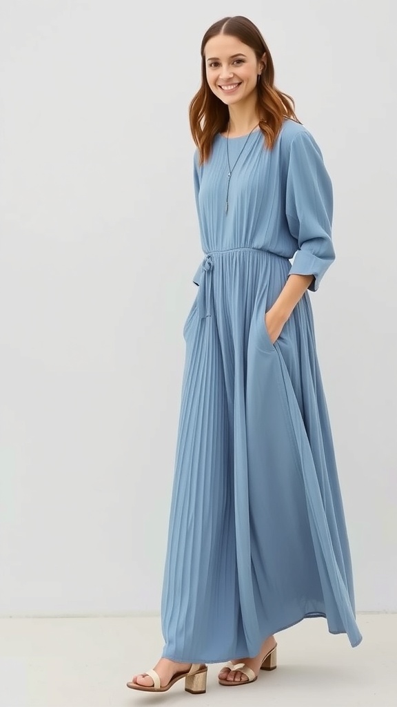 A woman wearing a dusty blue pleated long sleeve maxi dress with a cinched waist and pockets, smiling while standing against a light background.