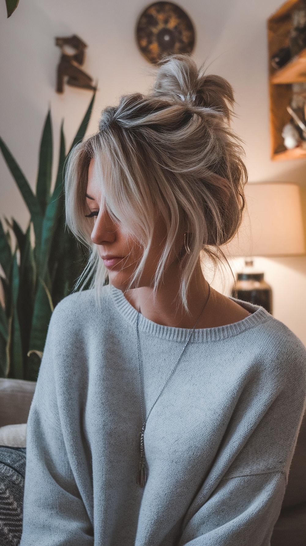 A woman with a messy bun and face-framing layers, wearing a cozy sweater, with plants in the background.