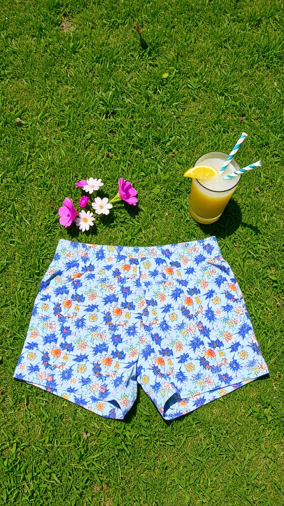 A pair of stylish blue and white checkered shorts on green grass, next to a drink and flowers.